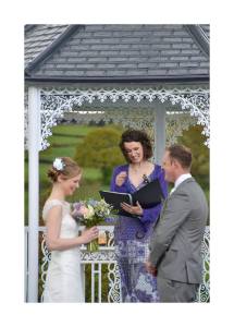 A bride and groom stand in front of a permanent gazebo smiling and laughing whilst their celebrant Keli Tomlin officiates their wedding ceremony. The wind is blowing. The bride is laughing.