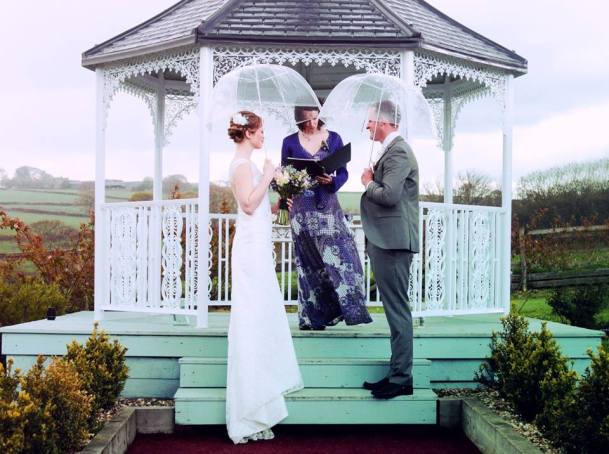 A bride and and groom stand beneath clear umbrellas on the first step of a wooden gazebo. Their Celebrant Keli Tomlin stands beside them officiating over their outdoor wedding ceremony