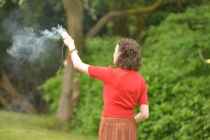 Pagan Celebrant Keli tomlin smudges the ceremony space in preparation for a Baby NAming. She is wearing a red cardigan and the smoke trails from her raised right hand.