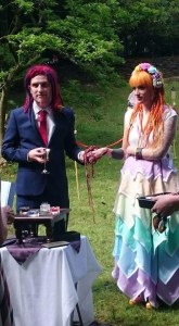 An Alternative Bride and Groom in rainbow colours stand together with their hands bound in Handfasting during their pagan wedding ceremony