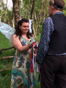 A Fairy Bride wearing wings and a white and green dress stands with her eyes closed smiling. Her hand is tied to her husbands with colourful handfasting cords by Celebrant Keli Tomlin