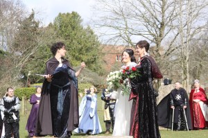 Pagan Celebrant Keli Tomlin stands in a blue dress and brown cape turning towards two Brides dressed in Medieval Wedding Dresses