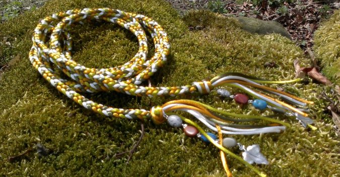 a handfasting cord made from gold, white and green cords lies curcled on a patch of moss. Semi-precious stones and silver ivy pendants are attached to each end.