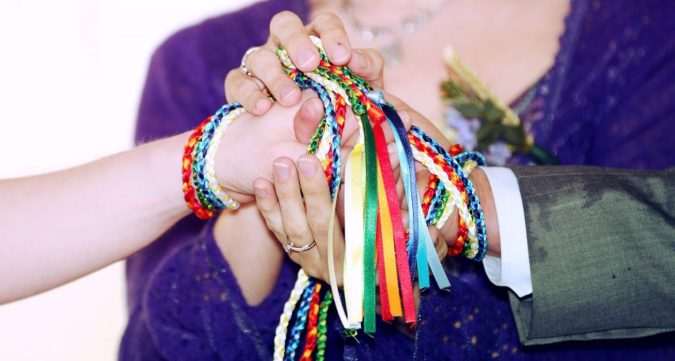 a couples hands are bound together with rainbow coloured handfasting cords. The Celebrant, Keli Tomlin, holds these joined hands between her own.