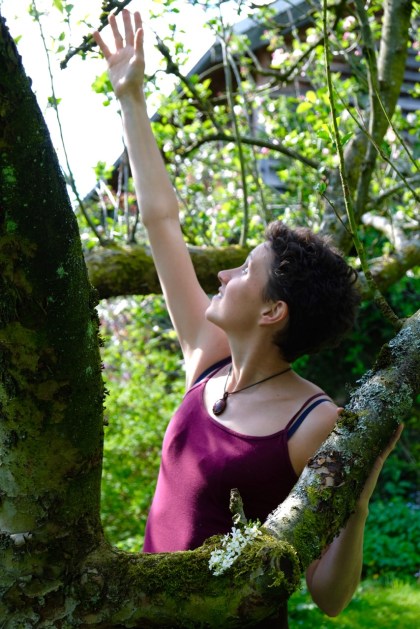 A woman stands beside the branches of a tree and reaches up with one hand towards the sky