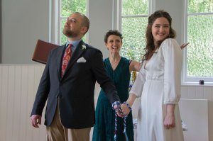A Bride and Groom have their hands tied as part of a handfasting ritual. They are smiling at their guests. The Celebrant Keli Tomlin stands behind them smiling, her arms spread to present them to the guests.