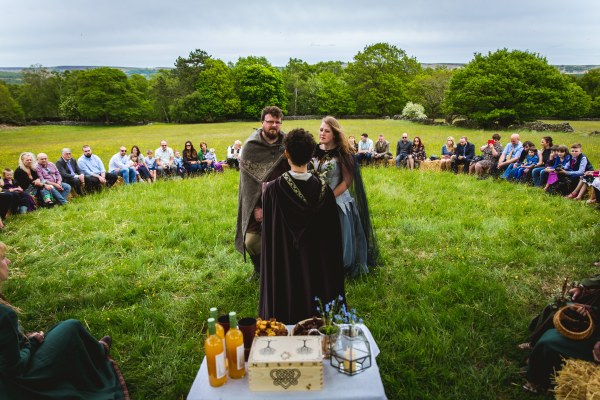 A Viking Bride and Groom stand at the centre of a circle of friends and family during their wedding ceremony led by Celebrant Keli Tomlin. There is an altar in the foreground with a wooden box, candles, forget-me-nots, bottles of mead and cake.