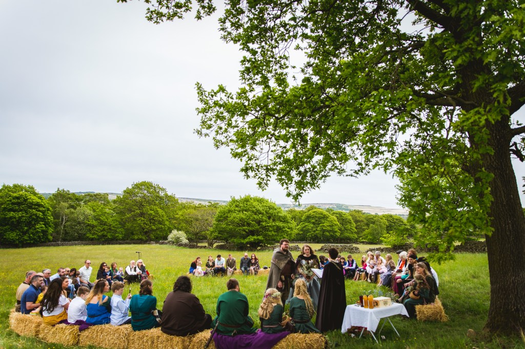 A Pagan Viking Wedding is conducted beneath the branches of an overhanging Oak tree. Bride, Groom and Celebrant Keli Tomlin are dressed in Viking attire at the centre. Guests sit on hay bales in a circle around them