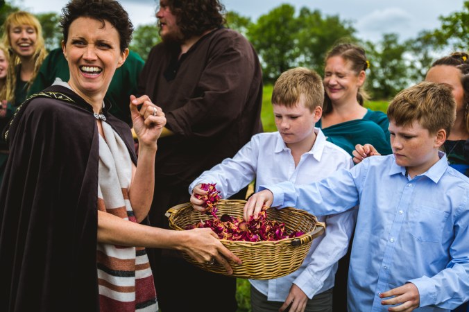 Keli Tomlin Celebrant at a Viking Wedding Ceremony hands dried pink rose petal confetti to two young boys. She is looking over her shoulder and smiling broadly