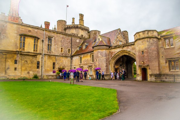 The Outside of Thornbury Castle, a yellow stone building with turrets and an archway, with a lawn in front. Wedding guests move towards the entrance carrying purple umberellas