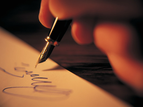 the tip of a fountain pen hovers above a page where a looping signature has been made in blue ink