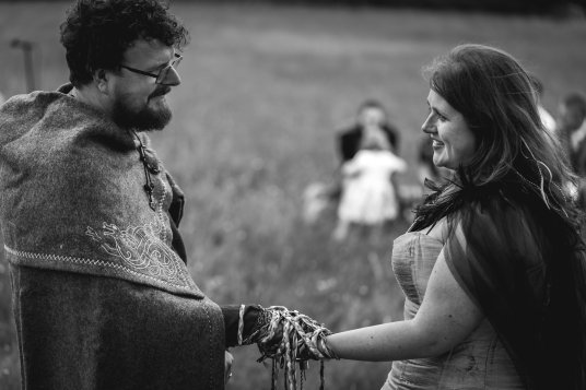 Bride and Groom in Viking clothes stand together and smile at one another. Their hands are bound with Handfasting cords by Keli Tomlin. The image is in black and white.