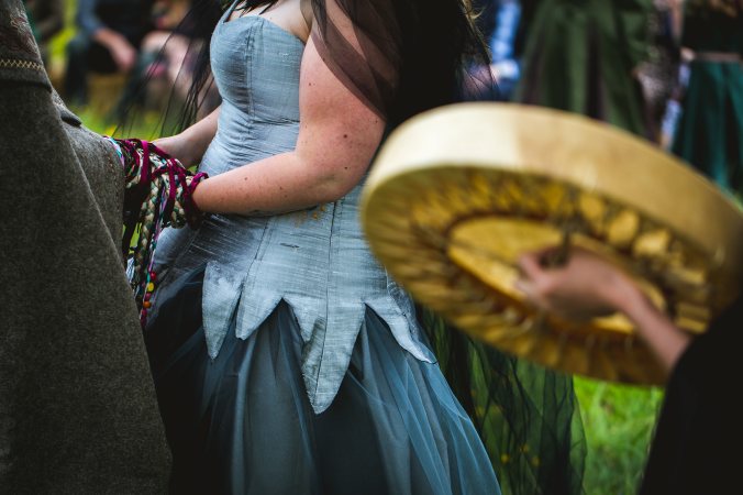 the hands of a couple are bound together with many coloured cords and ribbons, in a Handfasting. In the foreground a drum is visible in the hands of Celebrant Keli Tomlin