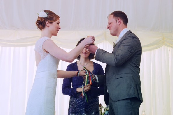 a bride and groom stand together holding a cup. Their other hands are bound with rainbow handfasting cords. their Celebrant Keli Tomlin stands behind them smiling.