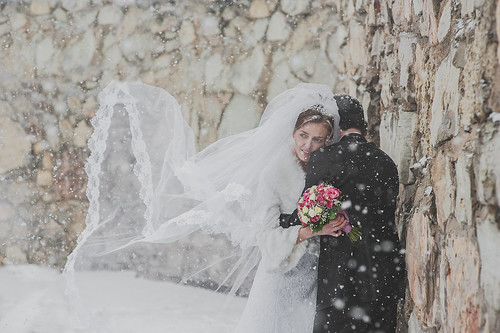 A Bride and Groom stand outside next to a tall stone wall. The groom's back is to the camera, the bride is embracing him and peering over his shoulder to the distance. Her veil is being blown out beside her by the wind. There is snow falling.