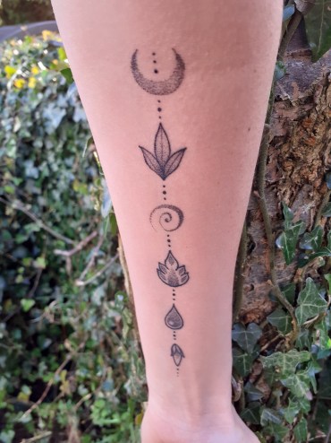 An inner arm tattoo with six symbols including one for each of the four elements - earth, air, fire, water - and a crescent moon. Behnd the arm is a tree covered in ivy.