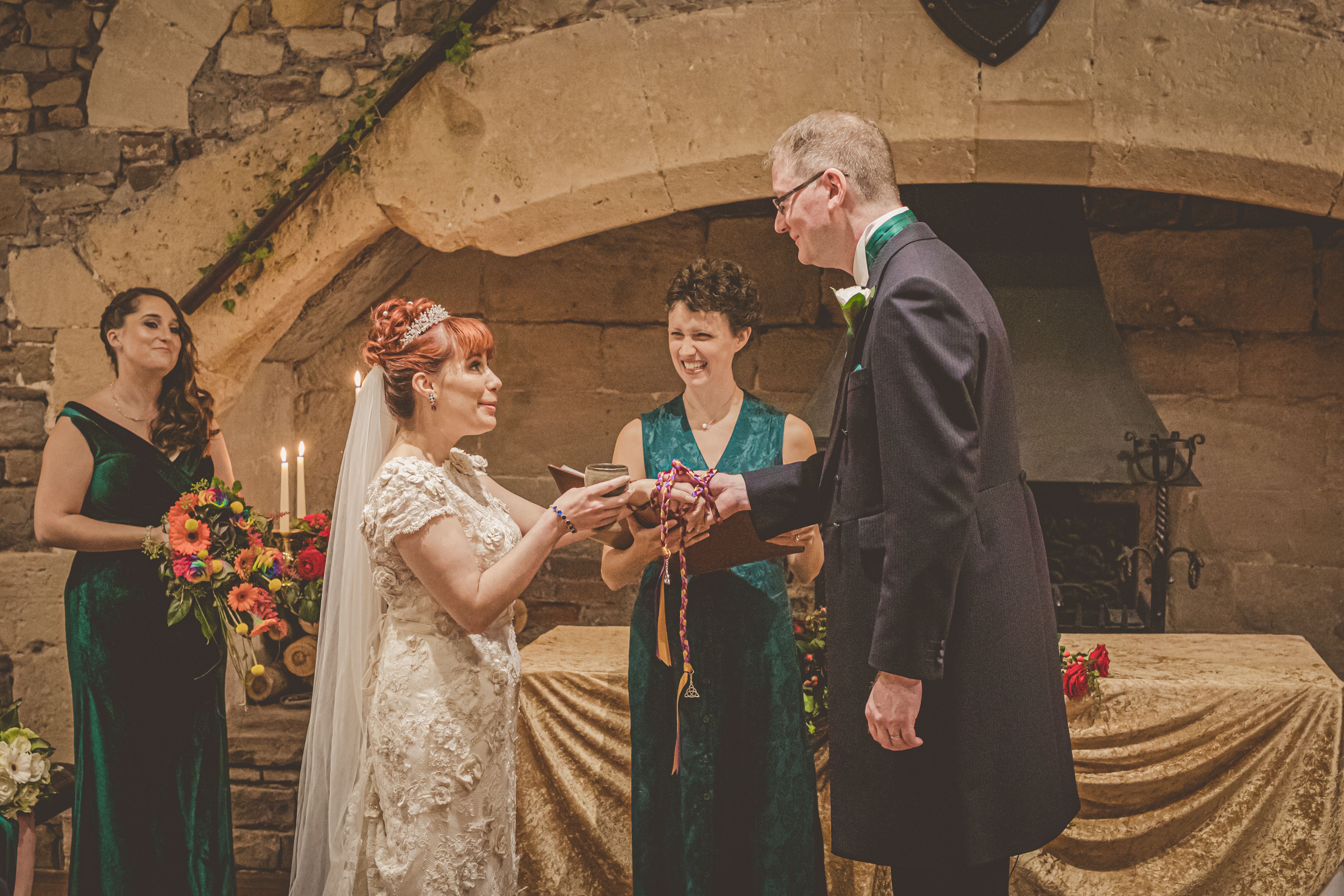 A Bride and Groom stand before a large stone fireplace. The Celebrant Keli Tomlin is standing behind them, laughing at something the bride has said. The couple's hands are tied with a purple and gold handfasting cord.
