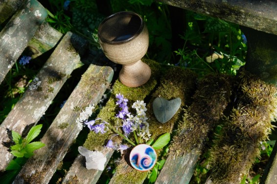 A stone chalice filled with water stands on a mossy old bench, beside it is a heart shaped stone, a ceramic pendant with a swirl motif, a piece of quartz crystal and a small bunch of flowers. A small altar by Keli Tomlin