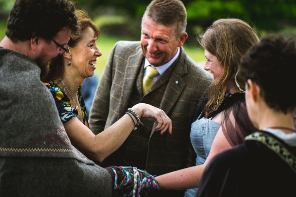 A man and woman laugh with the bride and groom who are in the process of being handfasted; their hands are tied together with many colourful ribbons. Part of a Handfasting by Keli Tomlin Ceremonies