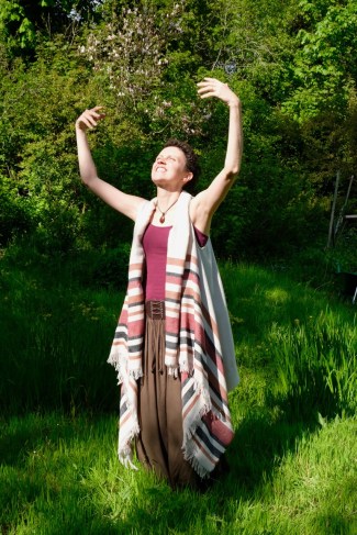 a woman, keli tomlin, stands amongst trees and grass, wearing a long green skirt, purple vest and long white jacket. her arms are raised to the sky and her face turned upward. she is smiling at the sun.