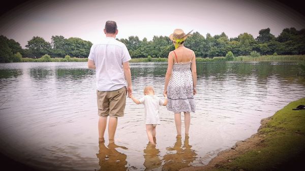 a man, woman and small child stand barefoot in the water of a lake with their backs to the camera. the child is in the middle holding the adults' hands.