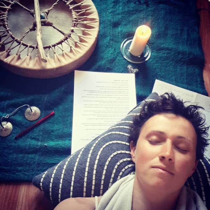 Keli, a woman with short dark hair, is laying with her head on a cushion and her eyes closed, in the bottom right corner. Behind her is a blue blanket and on top is a candle, a piece of paper, hand bells and a skin drum.