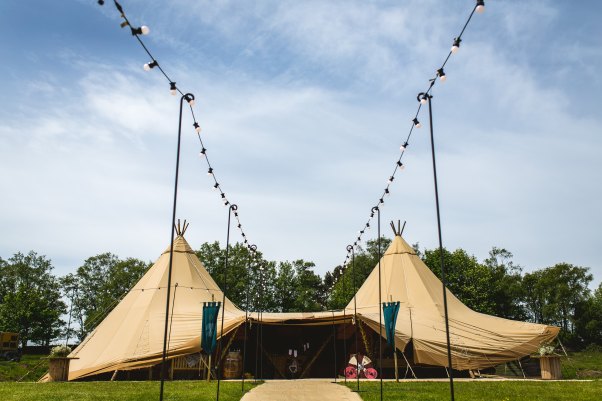 A tipi wedding tent set stands against a blue sky and green trees, there is a walkway with lights strung on either side leading to the entrance and green medieval banners fluttering beside